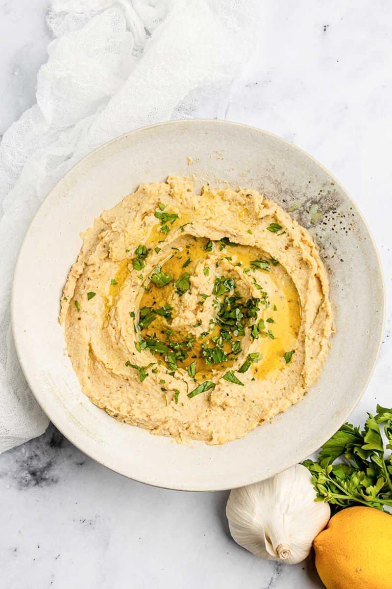 Overhead view of homemade garlic hummus, garlic bulb, lemon, and parsley.
