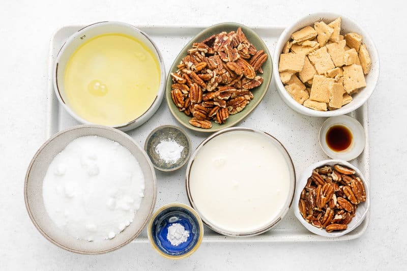 An overhead view of the ingredients for Luby's butternut brownie pie on a white tray. Pictured are bowls of egg whites, heavy cream, sugar, pecans, and broken graham crackers, along with smaller dishes of vanilla, baking powder, and cream of tartar.