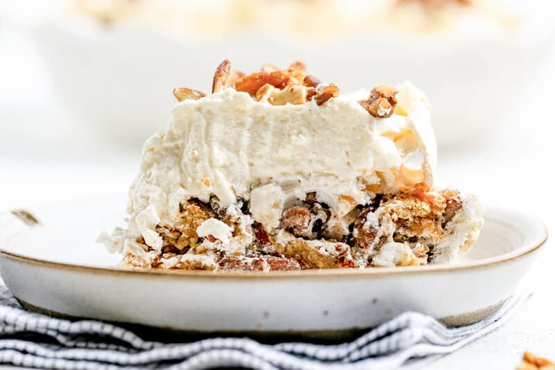 A side view of a slice of Luby's butternut brownie pie on a plate. The layers are distinct, showing the crunchy meringue and graham cracker base below a thick layer of whipped cream, all garnished with pecans