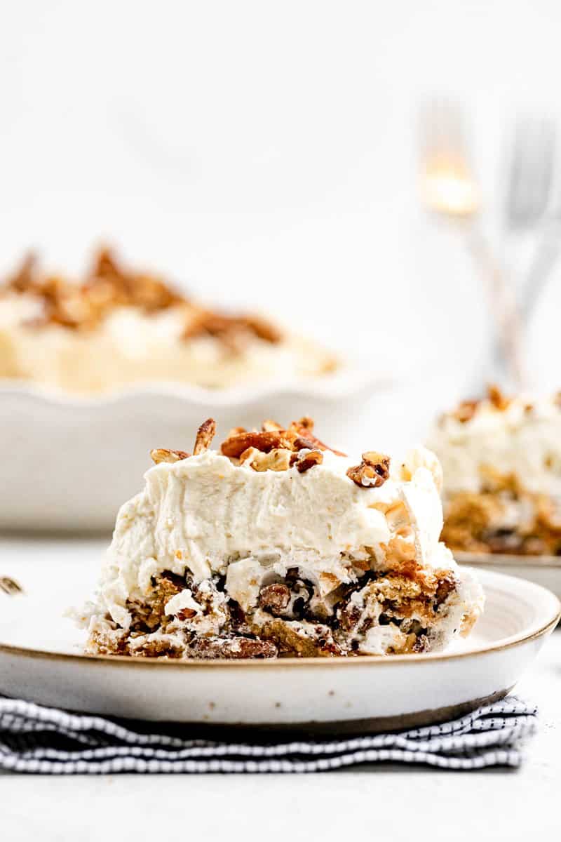 A slice of Luby's butternut brownie pie served on a plate in the foreground. The full pie, topped with whipped cream and pecans, is visible in the blurred background.