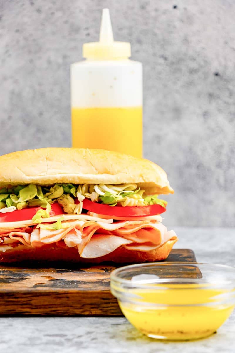 Copycat Mr. Submarine sub sauce in a small bowl and squeeze bottle and a sub sandwich.