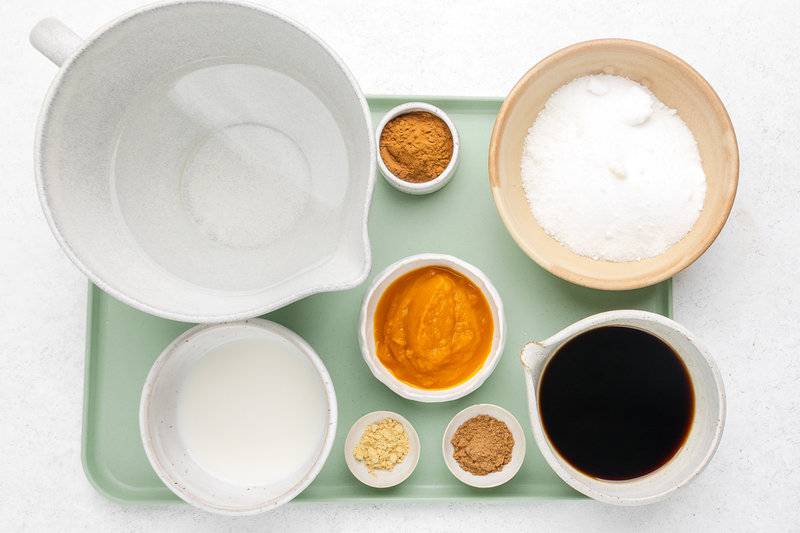 An overhead view of the ingredients for a homemade pumpkin spice latte on a green tray. Pictured are bowls of water, strong coffee, milk, sugar, pumpkin puree, and smaller dishes of ground cinnamon, nutmeg, and ginger.