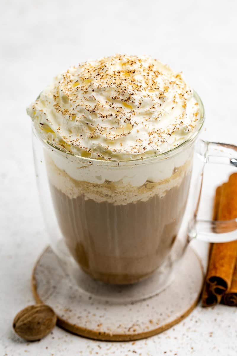 A detailed close-up of a pumpkin spice latte, focusing on the large swirl of whipped cream topping and the dusting of pumpkin pie spices. Whole cinnamon sticks and a nutmeg are visible beside the mug.