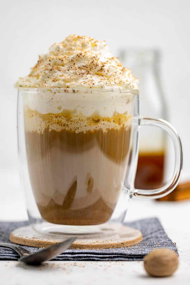 A homemade pumpkin spice latte in a double-walled glass mug, topped with whipped cream and spices. A bottle of pumpkin spice syrup and whole spices are visible in the background, creating a cozy autumn scene.