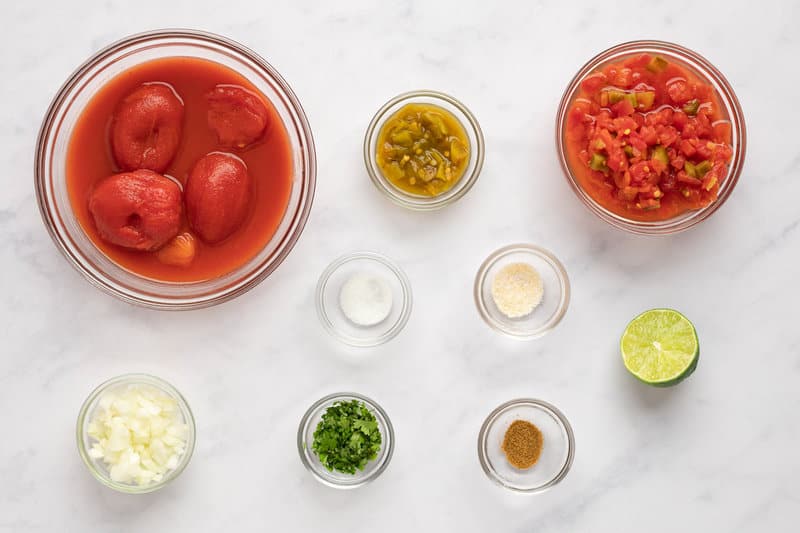 Overhead view of salsa ingredients in glass bowls: whole tomatoes, diced tomatoes, jalapenos, onion, spices, cilantro, and a lime.