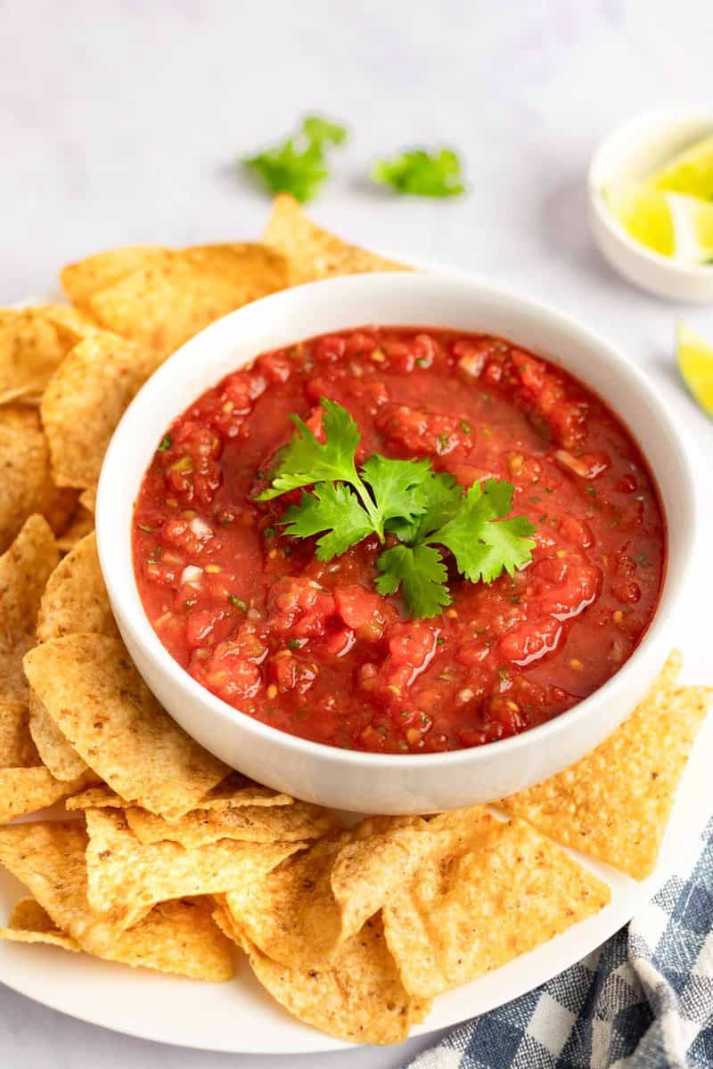 An overhead shot of a white bowl of red salsa, garnished with cilantro, surrounded by tortilla chips on a white plate.