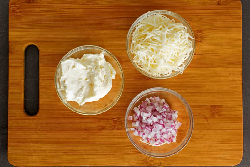 An overhead shot of the three simple ingredients for Jarlsberg cheese dip arranged on a wooden cutting board. Pictured are small glass bowls containing shredded Jarlsberg cheese, mayonnaise, and finely diced red onion.