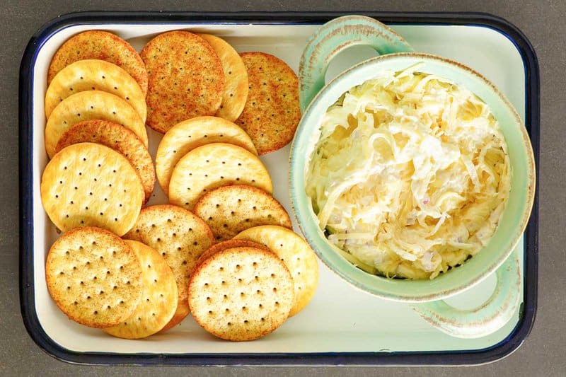A serving tray with a light green bowl of Jarlsberg cheese dip and an assortment of round crackers. The creamy, cheesy dip is ready to be served as an appetizer.