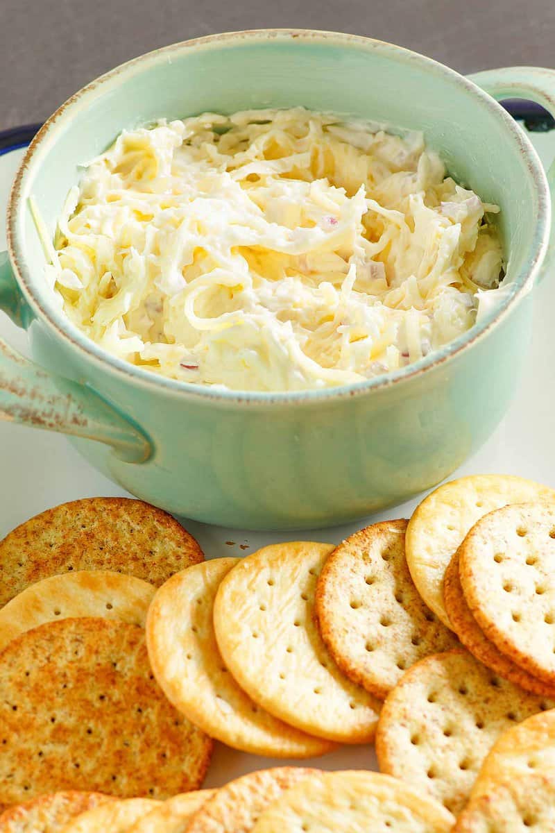 A close-up of a green bowl filled with creamy Jarlsberg cheese dip, highlighting its texture of shredded cheese and finely diced red onion. A fan of assorted crackers is arranged in front of the bowl.