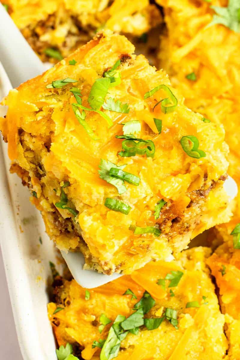A spatula lifting a cheesy slice of Mexican cornbread casserole from a white baking dish.