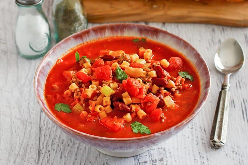 A rustic bowl filled with hearty, homemade Pasta e Fagioli soup. The rich tomato broth is brimming with ground beef, pasta, beans, and vegetables, and is garnished with fresh parsley.