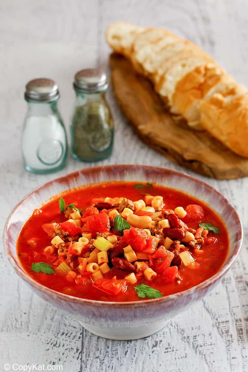 A welcoming bowl of Pasta e Fagioli soup, served with a loaf of crusty bread in the background. The hearty soup is filled with meat, beans, pasta, and vegetables in a rich tomato broth.