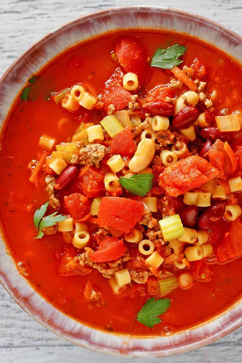 A close-up, overhead view of a bowl of Olive Garden's Pasta e Fagioli soup. The image focuses on the rich texture and vibrant colors of the ground beef, beans, pasta, and vegetables in the tomato broth.