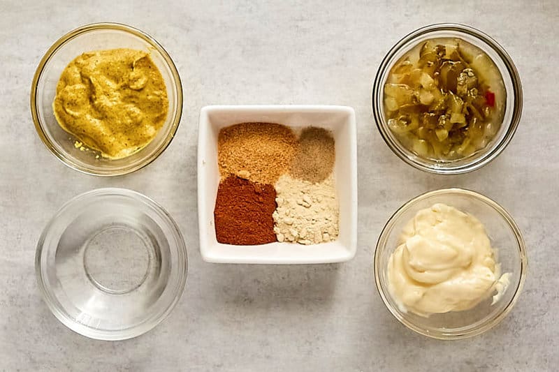 An overhead shot of the ingredients for copycat Big Mac sauce. Glass bowls hold mayonnaise, India relish, brown mustard, and white vinegar, while a square dish contains mounds of paprika, onion powder, garlic powder, and other seasonings.