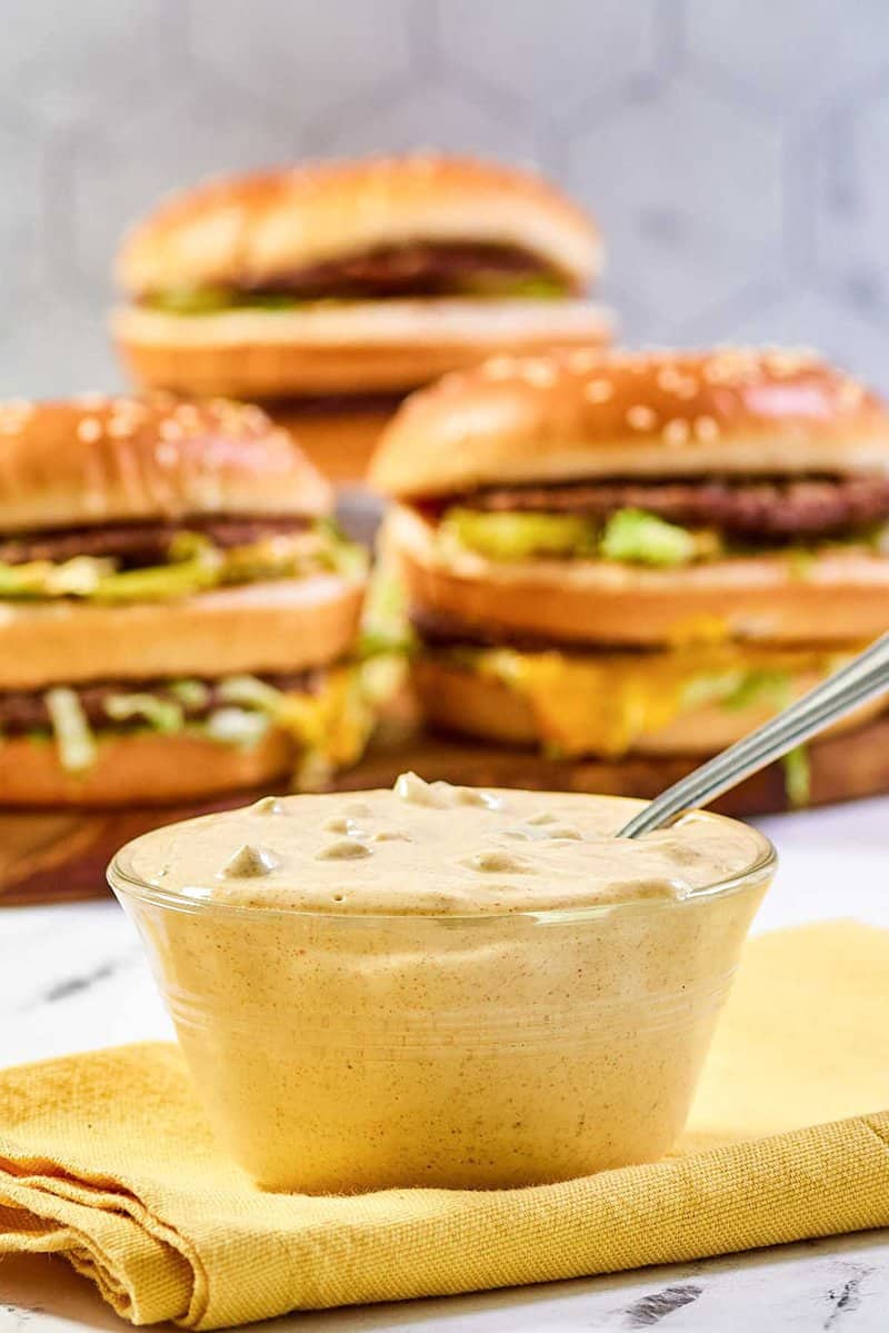 An inviting shot of a glass bowl of homemade Big Mac sauce resting on a yellow napkin. The creamy sauce is in sharp focus in the foreground, while three delicious-looking homemade Big Mac burgers are staged in the soft-focus background.