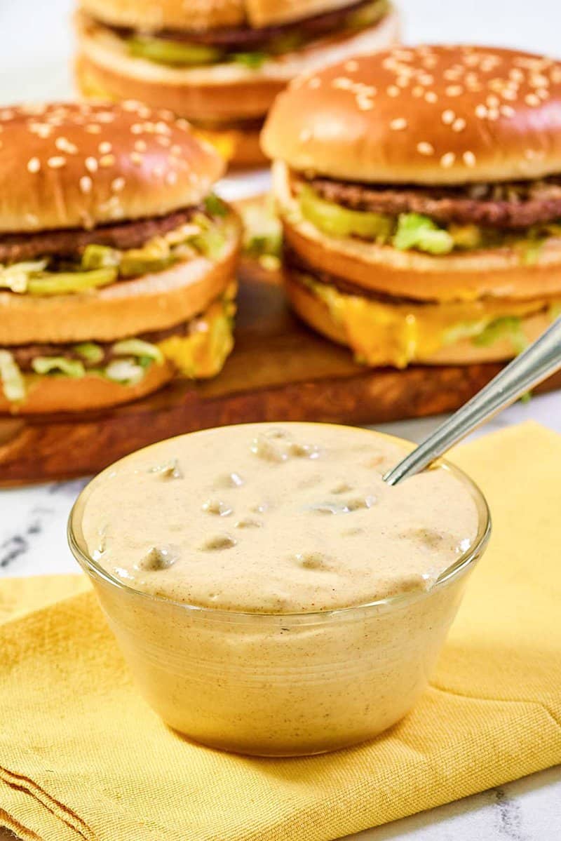 A close-up shot of a glass bowl filled with creamy homemade Big Mac sauce, with a spoon dipped inside. The sauce shows visible texture from the relish, and homemade Big Mac burgers are artfully arranged in the blurred background.