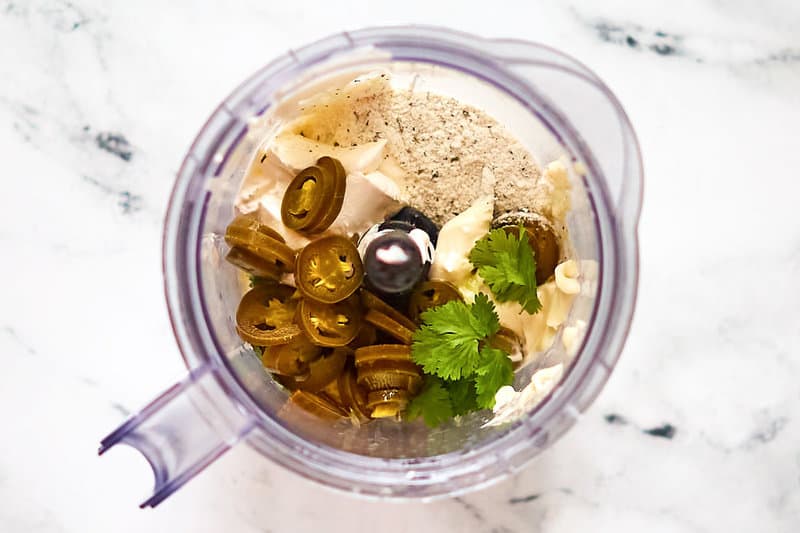 An overhead view looking into a blender pitcher filled with the ingredients for the creamy jalapeno dip. Pickled jalapeno slices, cilantro, sour cream, mayonnaise, and dry ranch dressing mix are visible, ready to be blended into a smooth sauce.