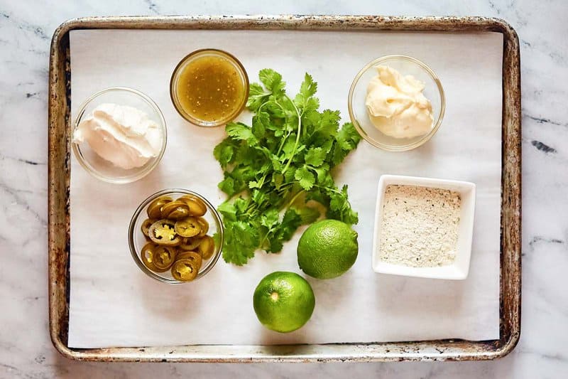 Overhead view of dip ingredients: sour cream, mayo, salsa verde, jalapenos, limes, cilantro, and ranch seasoning on a tray.