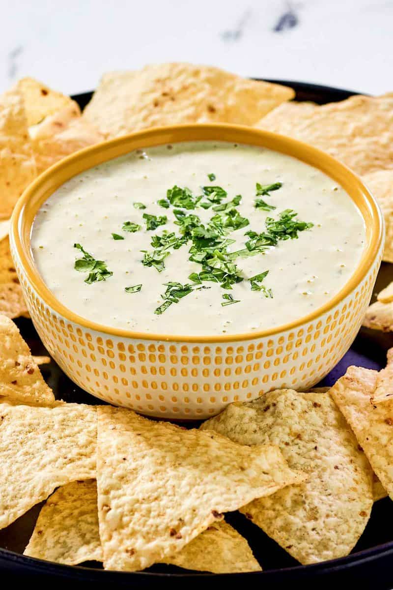 A close-up, angled shot of a textured yellow bowl filled with creamy jalapeno dip and garnished with fresh cilantro. The bowl is nestled in a bed of tortilla chips, highlighting the dip's smooth, inviting texture.