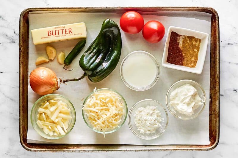 An overhead view of the ingredients for Chipotle queso blanco on a baking sheet. Pictured are fresh onion, poblano and serrano peppers, tomatoes, and garlic, along with bowls of shredded cheeses, cream, sour cream, and spices.