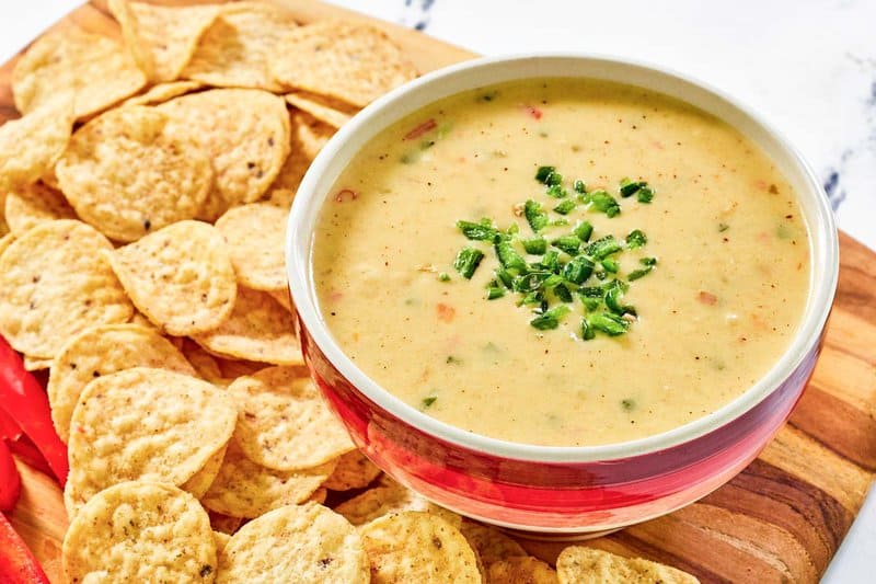 A bowl of creamy, homemade Chipotle queso blanco, garnished with diced jalapeños, is served on a wooden board alongside a generous pile of round tortilla chips.