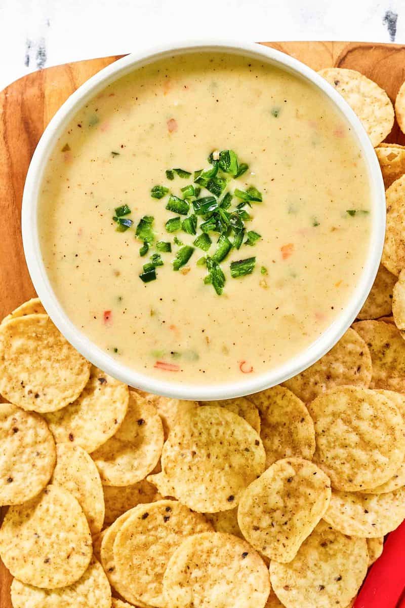 An overhead shot of a white bowl of Chipotle queso blanco, garnished with fresh diced peppers. The bowl is nestled in a bed of round tortilla chips, ready for dipping.