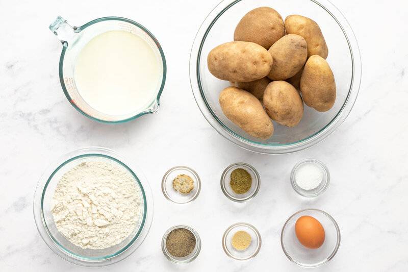 Overhead view of ingredients: russet potatoes, milk, flour, egg, and spices arranged on a white table.