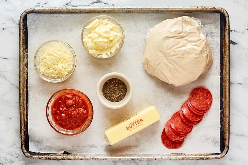 An overhead view of the ingredients for Little Caesars Crazy Puffs on a baking sheet. Pictured are a bag of pizza dough, pepperoni slices, a stick of butter, and bowls of pizza sauce, shredded cheese, and Italian seasoning.