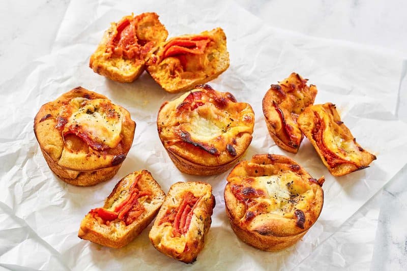 Several freshly baked Little Caesars Crazy Puffs are scattered on white parchment paper. Some of the muffin-sized pizza bites are whole, and others are cut in half to show the cheesy pepperoni filling.