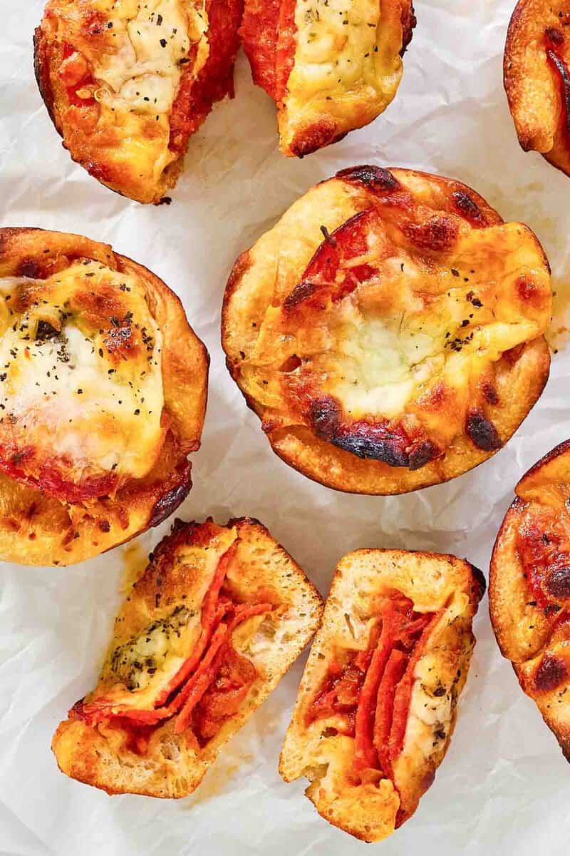 A close-up of several homemade Crazy Puffs on parchment paper. The image highlights the golden-brown, cheesy tops of the pizza muffins, with one cut in half to reveal the layered pepperoni inside.