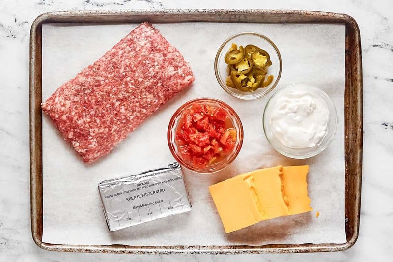 An overhead shot of the ingredients for Rotel sausage dip on a parchment-lined baking sheet. Pictured are a block of raw ground sausage, cream cheese, sliced Velveeta cheese, and bowls of Rotel tomatoes, sour cream, and pickled jalapenos.