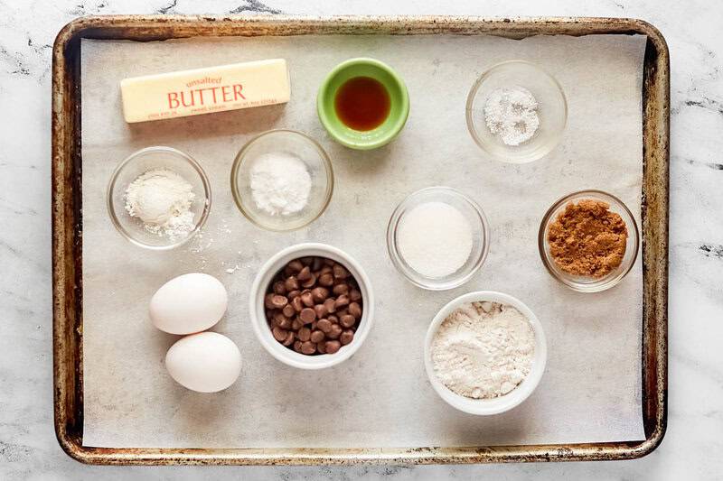 An overhead view of the ingredients for copycat Crumbl chocolate chip cookies on a baking sheet. Pictured are a stick of butter, eggs, and bowls of flour, sugars, cornstarch, vanilla, and milk chocolate chips.