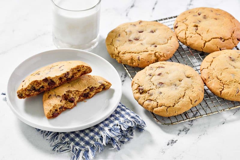 Freshly baked copycat Crumbl chocolate chip cookies are cooling on a wire rack. One cookie is broken in half on a plate to reveal its chewy, chocolate-filled center, served with a glass of milk.