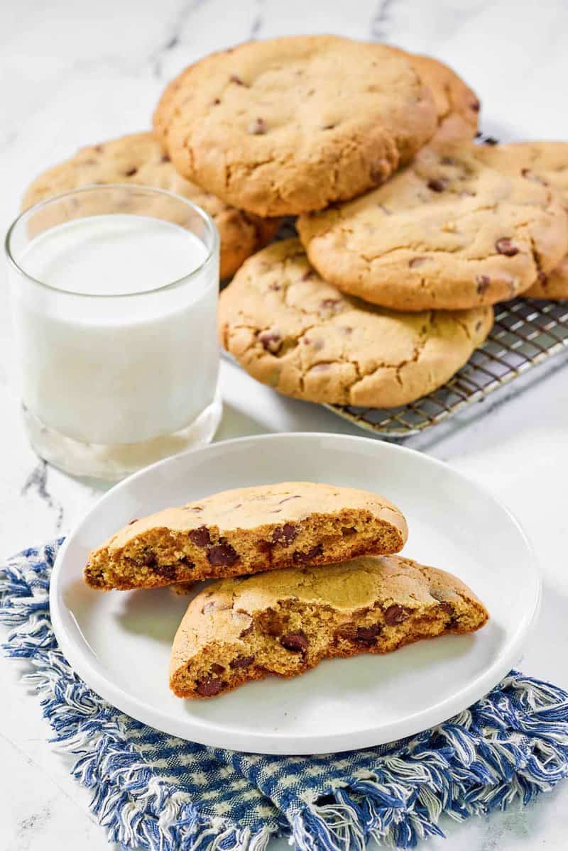 Freshly baked copycat Crumbl chocolate chip cookies are cooling on a wire rack. One cookie is broken in half on a plate to reveal its chewy, chocolate-filled center, served with a glass of milk.