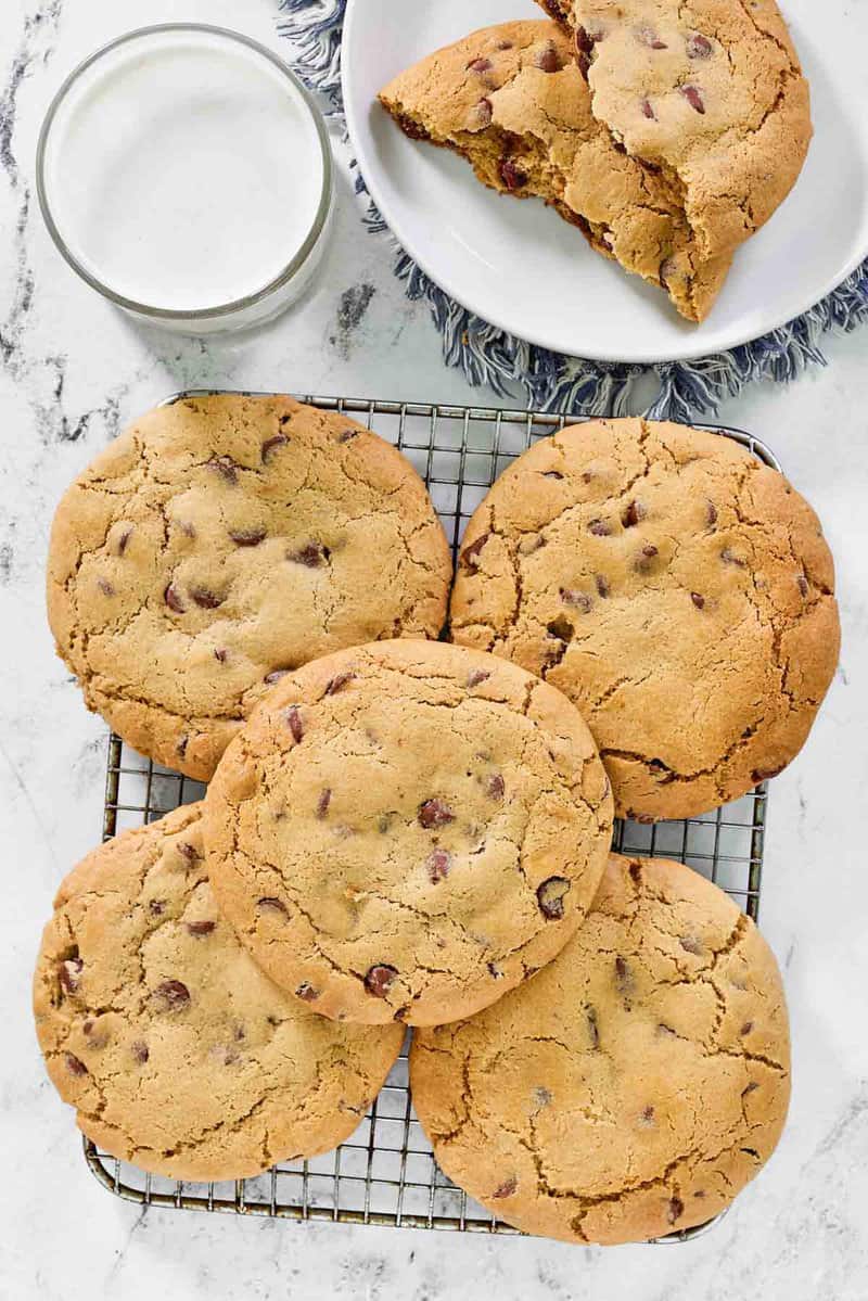An overhead, flat-lay view of several large, copycat Crumbl chocolate chip cookies on a wire cooling rack. A glass of milk and a plate with a cookie broken in half are placed nearby.
