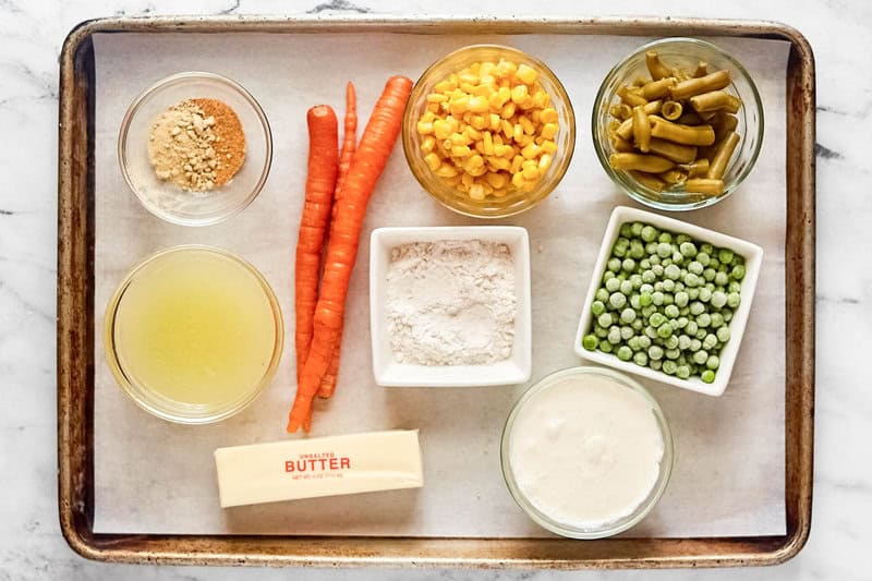 An overhead view of the ingredients for Dolly Parton's Dixie Stampede soup on a baking sheet. Pictured are carrots, butter, and bowls of chicken broth, cream, flour, peas, corn, green beans, and powdered seasonings.