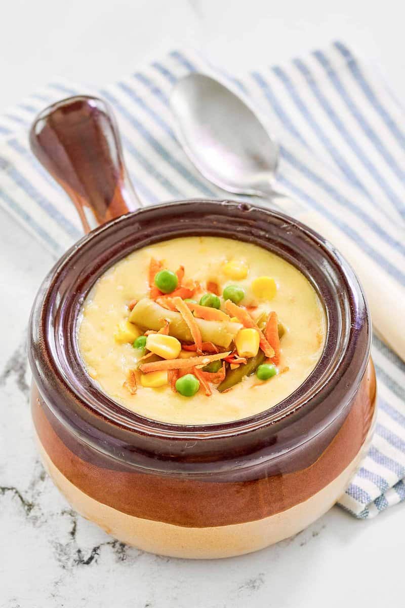 A close-up of a rustic brown crock filled with Dolly Parton's creamy vegetable soup. The image highlights the smooth, creamy texture and the colorful vegetable garnish.