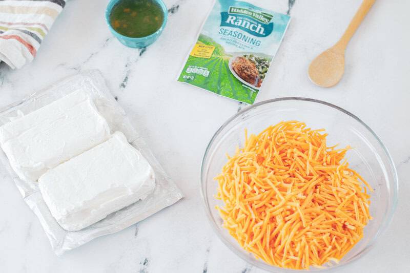 An overhead view of the ingredients for beer cheese dip. Pictured are two blocks of cream cheese, a packet of ranch seasoning, a bowl of shredded cheddar cheese, and a small bowl of beer.
