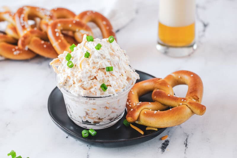 A bowl of cold beer cheese dip, garnished with green onions, is served with soft pretzels. A glass of beer is visible in the background, creating a perfect game day scene.