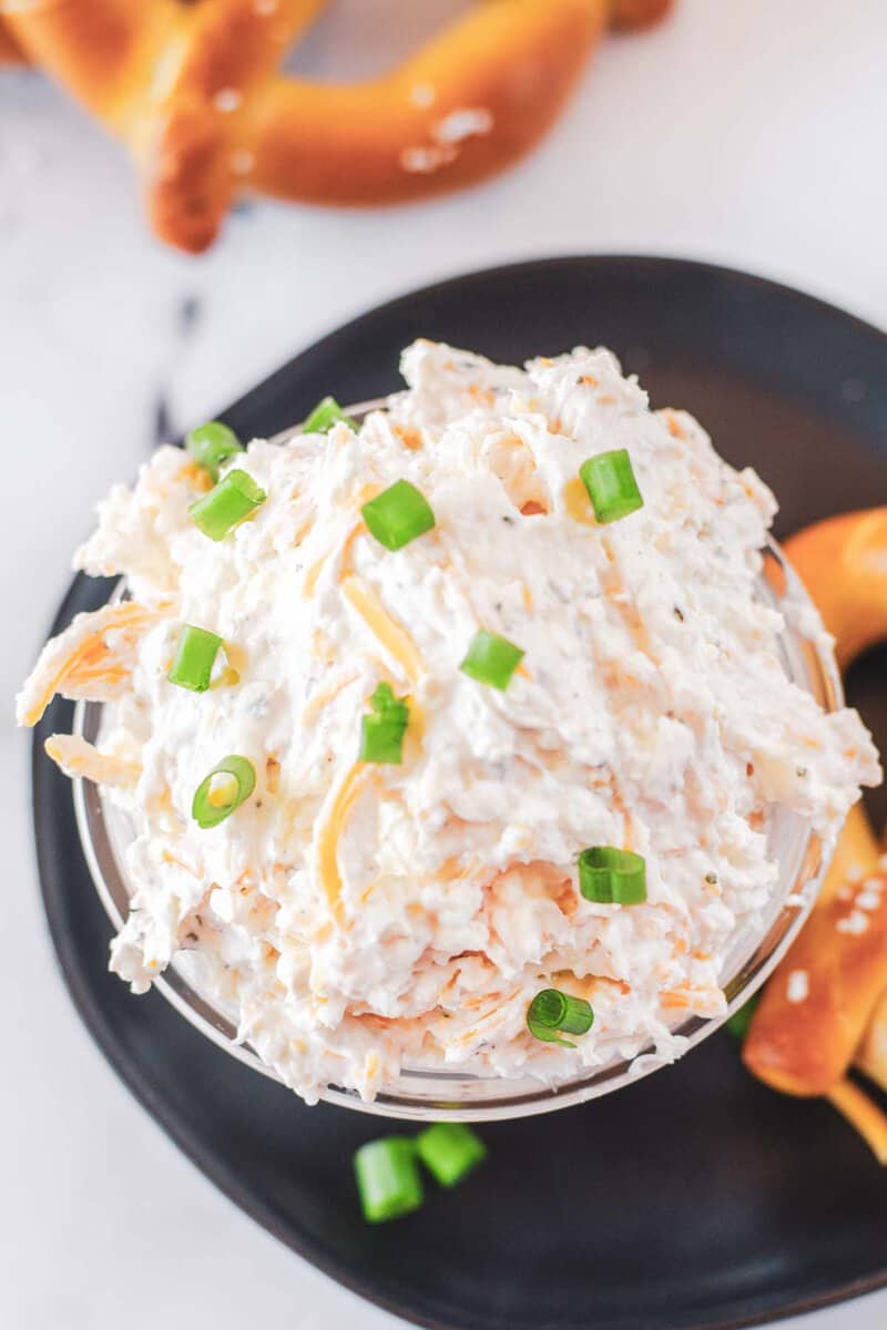 A close-up of a bowl of thick and creamy beer cheese dip, garnished with fresh green onions. A soft pretzel rests on the plate, with more pretzels visible in the soft-focus background.