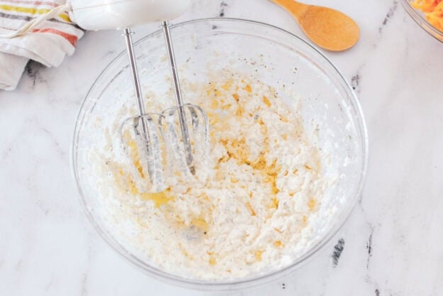 An electric hand mixer whips softened cream cheese, beer, and ranch seasoning together in a large glass bowl to create the creamy base for the beer cheese dip