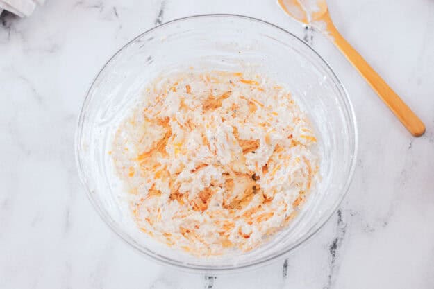 Shredded cheddar cheese is being folded into the cream cheese mixture in a glass bowl, creating the final texture for the cold beer cheese dip.