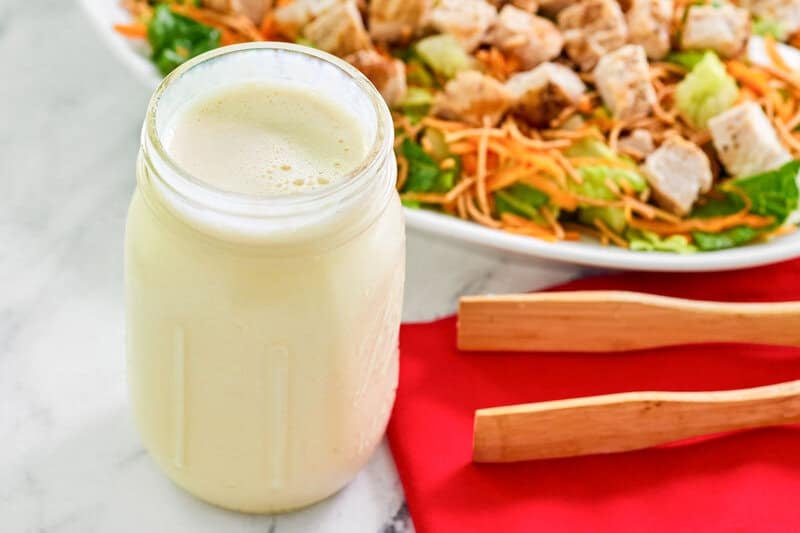 A glass jar filled with creamy, homemade Applebee's oriental salad dressing. In the blurred background, a large bowl of oriental chicken salad is visible, ready to be dressed.