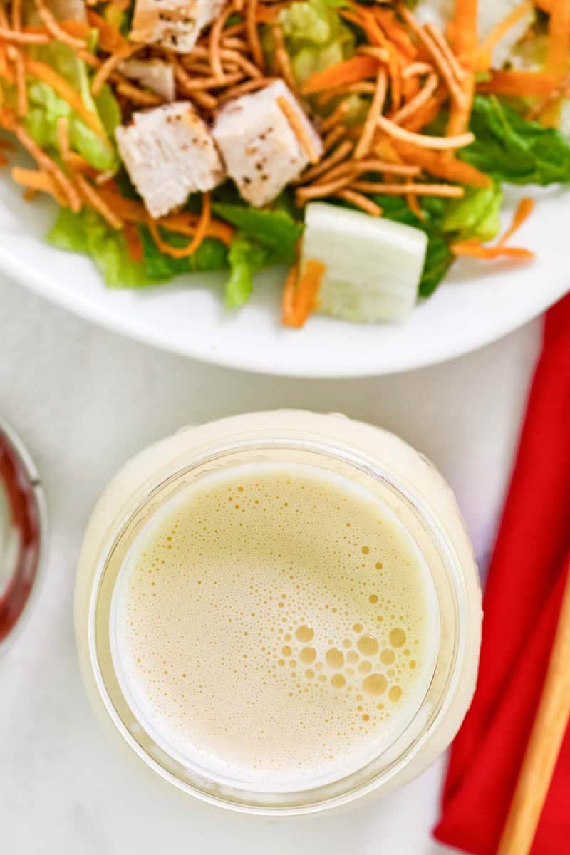 An overhead shot looking down into a glass jar of homemade oriental salad dressing. The creamy, slightly bubbly surface of the dressing is in focus, with a fresh oriental chicken salad partially visible in the background.
