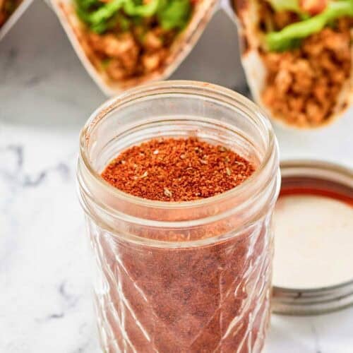 Homemade chicken taco seasoning in a jar and chicken tacos behind it.