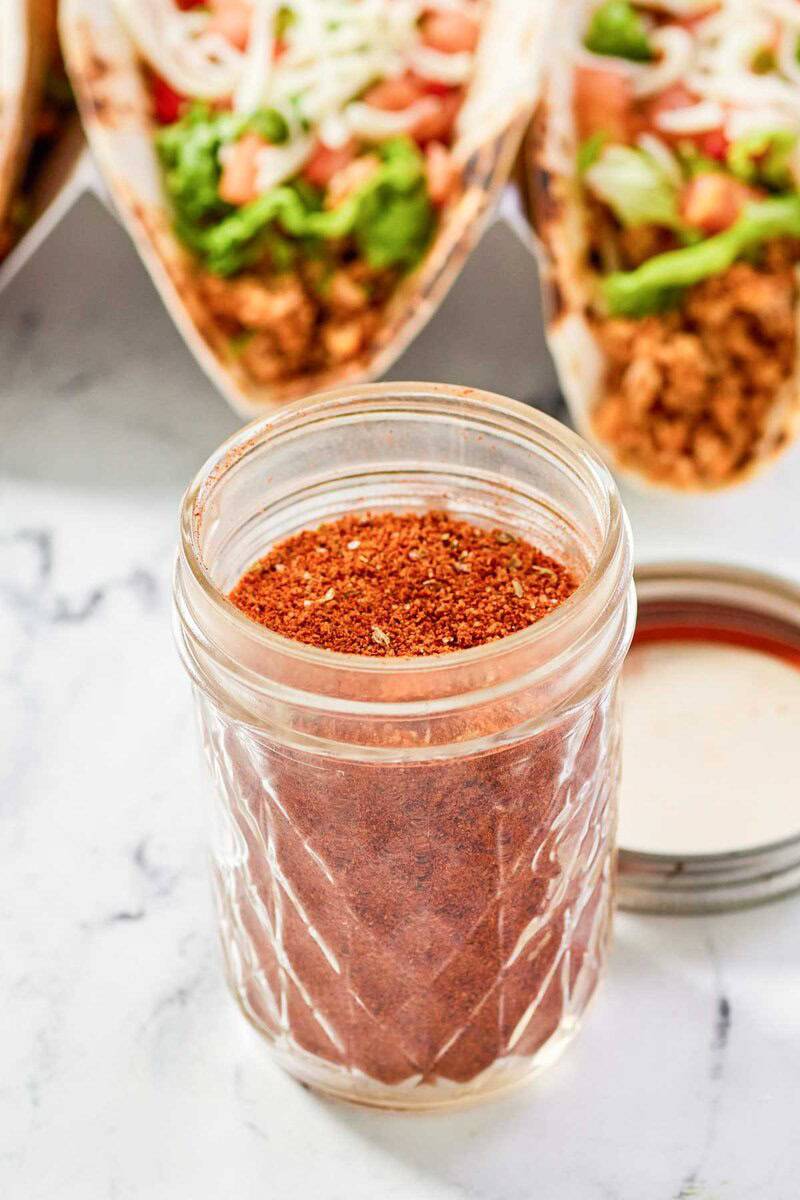Homemade chicken taco seasoning in a jar and chicken tacos behind it.