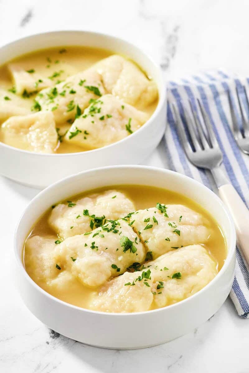 Two white bowls filled with flat dumplings in a golden broth, garnished with parsley, resting near forks on a blue striped napkin.