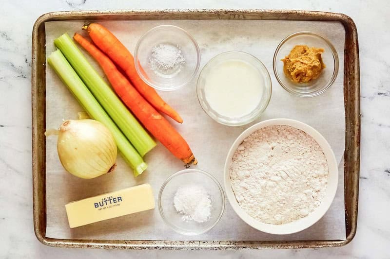 Ingredients on a tray: flour, milk, butter, chicken base, salt, onion, celery stalks, and carrots for making homemade dumplings.