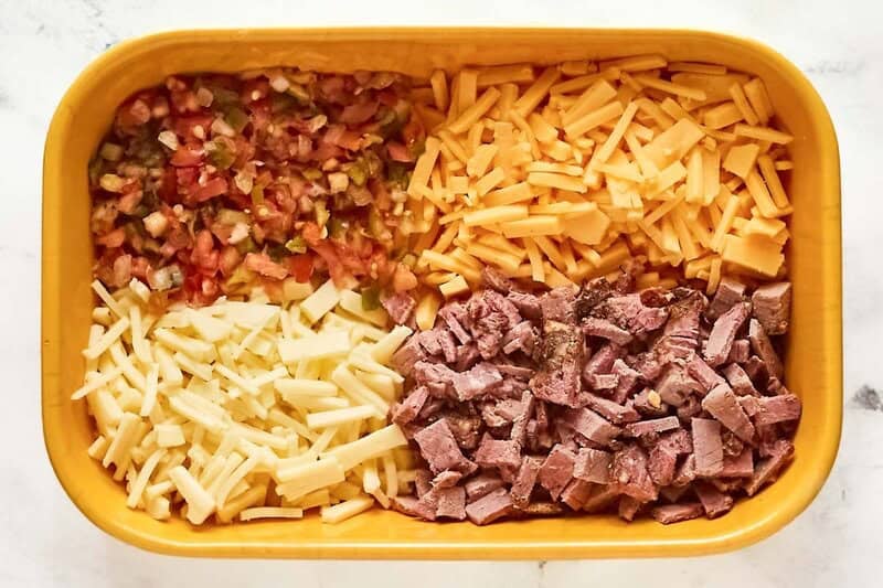 An overhead view of the unmixed ingredients for HEB brisket queso arranged in a yellow baking dish. Pictured are separate piles of shredded yellow cheese, shredded white cheese, chopped smoked brisket, and fresh pico de gallo.