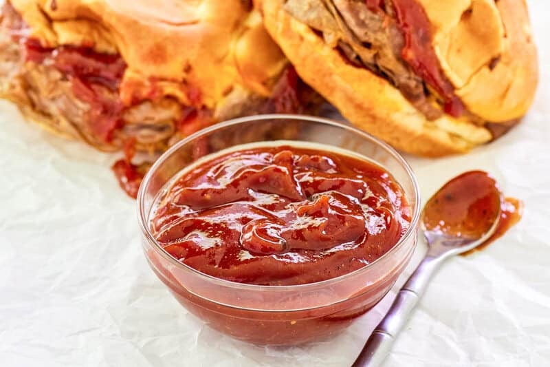 close-up of a small glass bowl filled with thick, tangy Arby's Red Ranch Sauce. In the blurred background, two roast beef sandwiches on onion buns are ready to be served with the sauce.